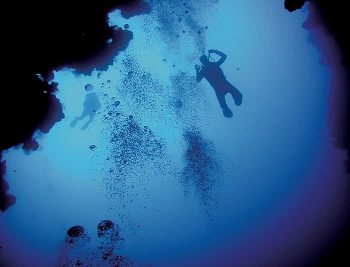 Two scuba divers are silhouetted against a dark cave opening, descending into the deep blue water. Bubbles rise around them as they explore the underwater cavern.