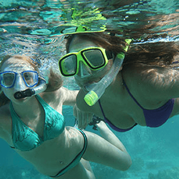 Two young ladies in snorkeling gear and goggles swim through the blue water.
