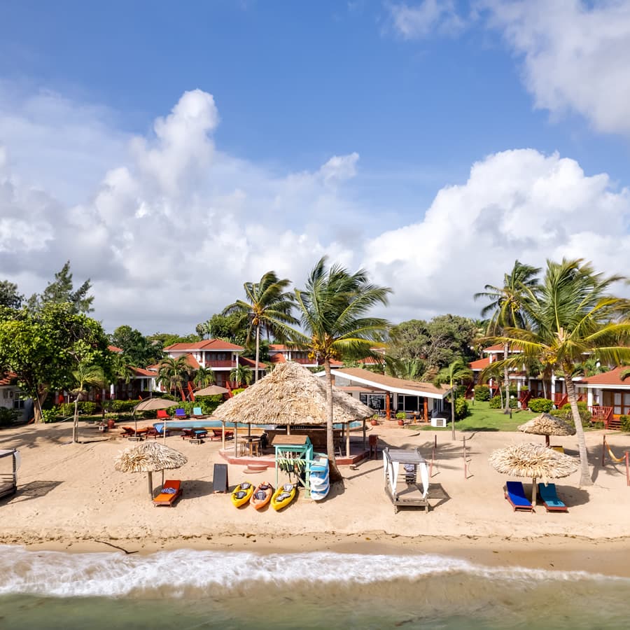 A tropical beach resort with a thatched-roof bar and restaurant. Kayaks are lined up on the shore, and beach umbrellas provide shade for lounge chairs. The resort has several buildings with colorful balconies overlooking the ocean.