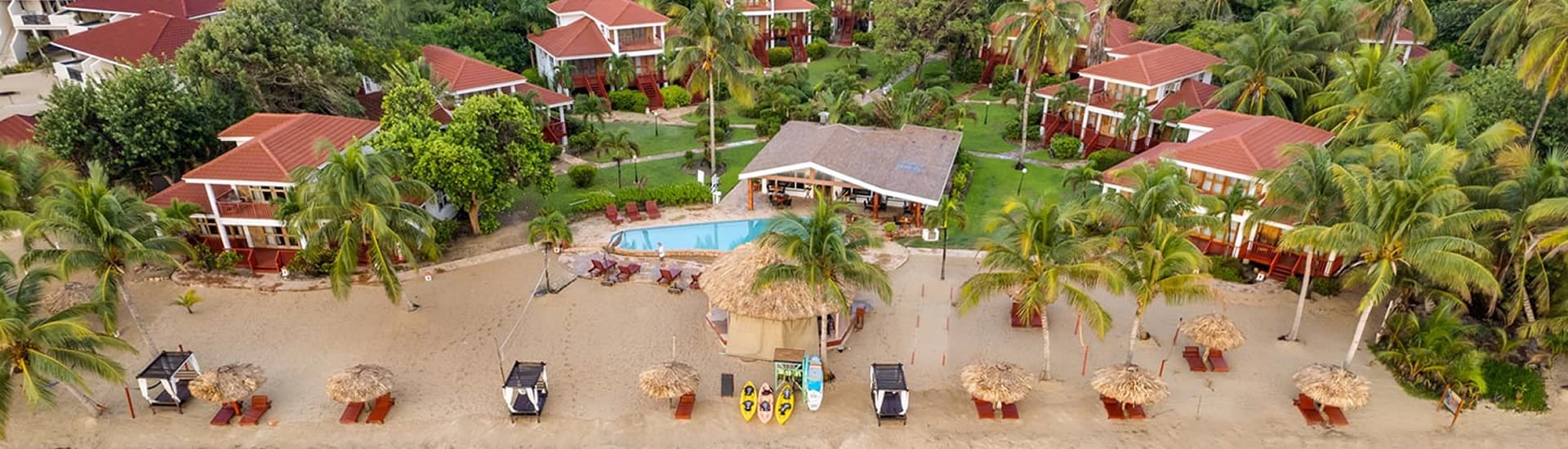 An aerial view of a tropical beach resort. The resort features multiple buildings with red roofs, a swimming pool, and a thatched-roof bar on the beach. Kayaks are lined up on the shore, and beach umbrellas provide shade for lounge chairs.