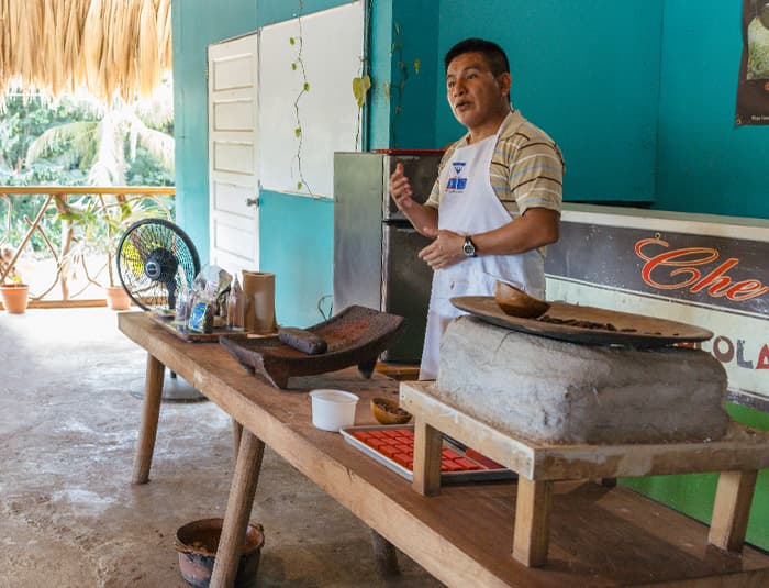 A teacher in a white apron shows a class how chocolate is made from the cacao bean.
