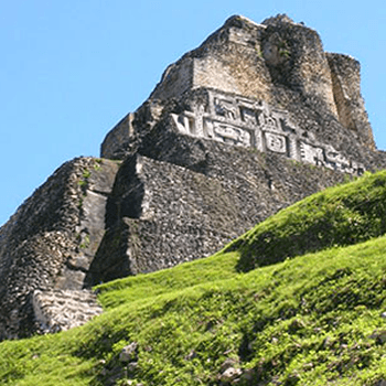 A steep, grassy slope leads up to a towering Mayan pyramid with intricate stone carvings on its facade. The sky is clear and blue.