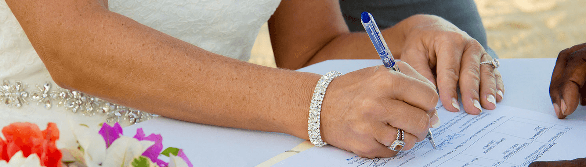 A bride in a white gown and diamond jewelry signs her marriage license on the beach.