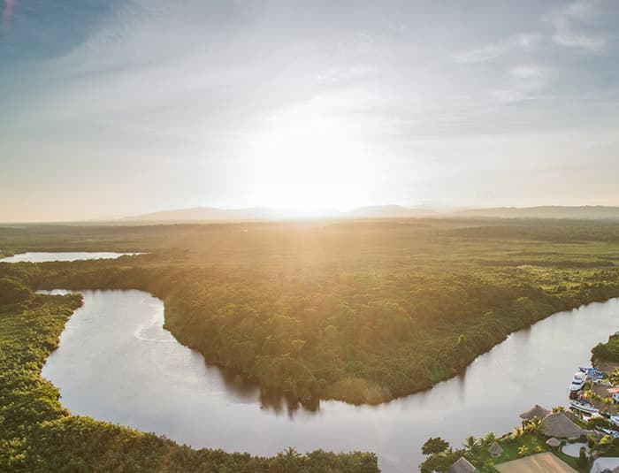 A beautiful aerial view of a river winding through a lush green landscape. The sun is setting in the distance, casting a warm glow over the scene. There are several small villages or towns visible along the riverbanks, with houses and boats scattered throughout. The water is calm and reflective, mirroring the sky and the surrounding vegetation.