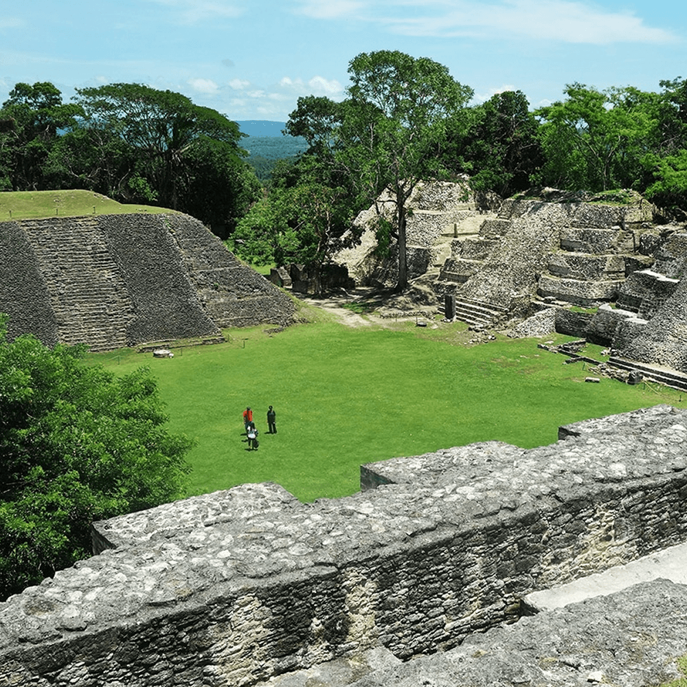 A panoramic view of the Xunantunich Mayan Ruins in Belize. The ruins are surrounded by lush green vegetation and include several stone structures, including a large pyramid with a staircase.