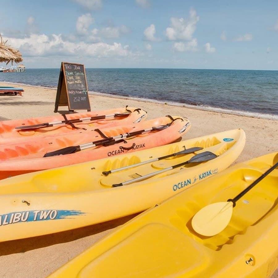 Several colorful kayaks lined up on a sandy beach. The kayaks are orange, yellow, and blue. A sign on the beach advertises "Happy Hour" with a discount. The ocean is visible in the background.