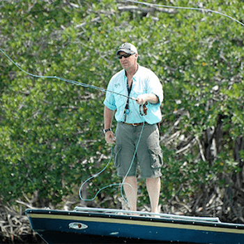 A large man in shorts stands on the bow of a boat, casting a fly-fishing line into the river.