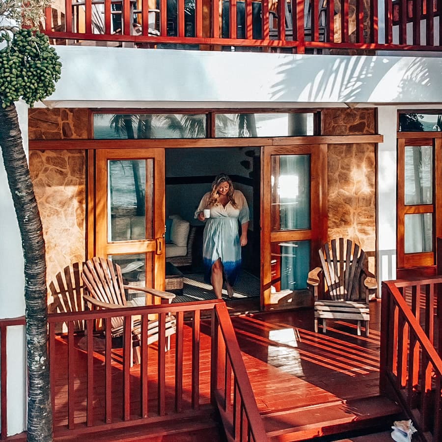 Woman in blue and white sun dress with cup in hand walking out of lodging accommodation on the wood porch. Stone walls in background.