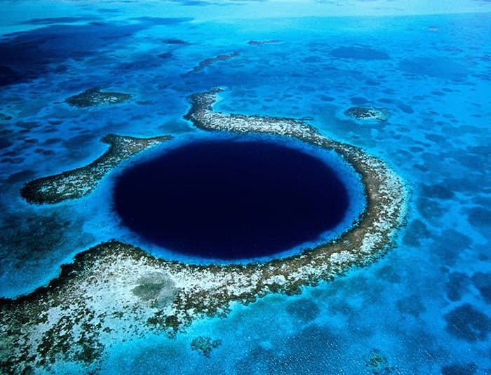 An aerial view of the Great Blue Hole in Belize. It's a circular, dark blue hole surrounded by a vibrant coral reef and turquoise ocean. The sky is clear with white clouds.