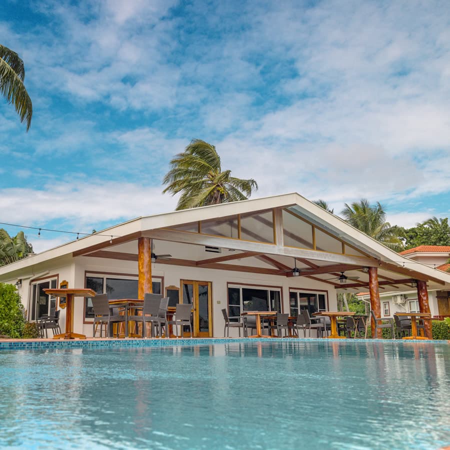 A beachfront restaurant with a large pool. The restaurant has a white exterior with a thatched roof and open-air seating. Palm trees line the property.
