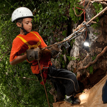 A young boy in a white helmet and orange shirt rappels carefully down a slope next to a waterfall.