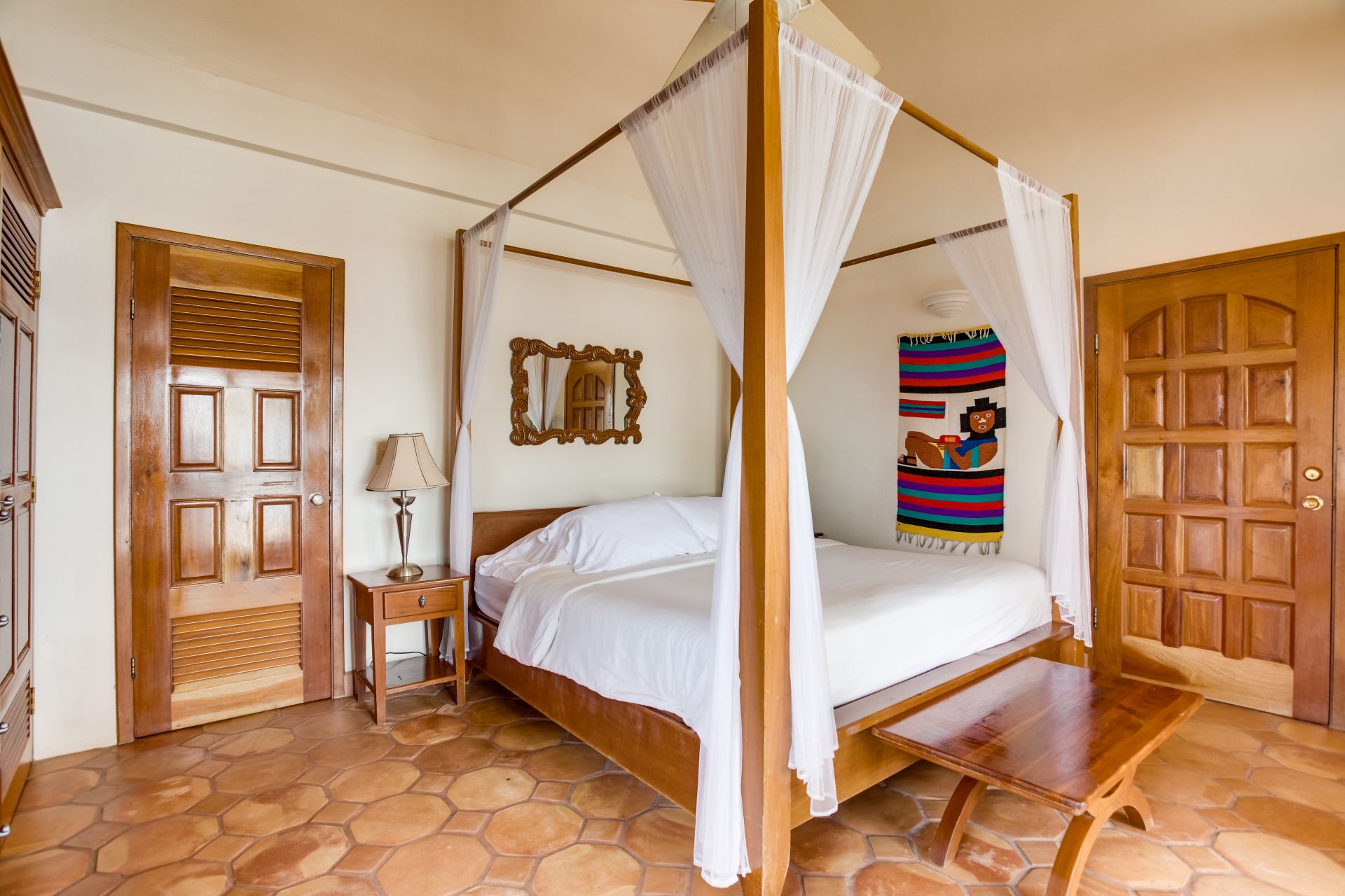 A spacious bedroom with a four-poster bed covered in white sheets. The bed has sheer white curtains hanging from the canopy. A colorful tapestry hangs on the wall behind the bed, and a wooden bench sits at the foot. The room has a tiled floor and a wooden nightstand with a lamp.