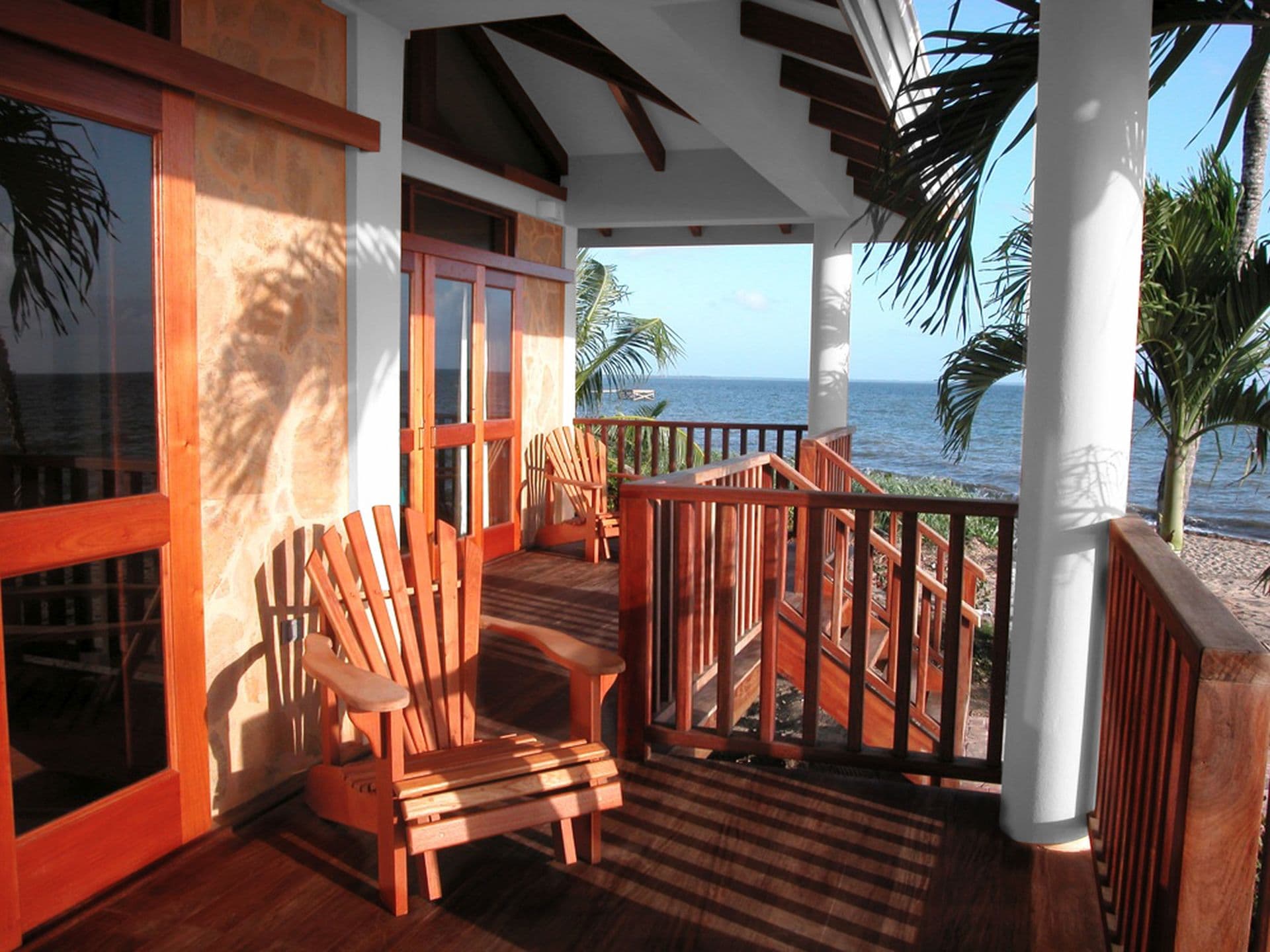 A spacious balcony with a wooden railing overlooking the ocean. A wooden Adirondack chair sits on the balcony, and there are stairs leading down to the beach. Palm trees are visible in the background. The sky is blue and sunny.