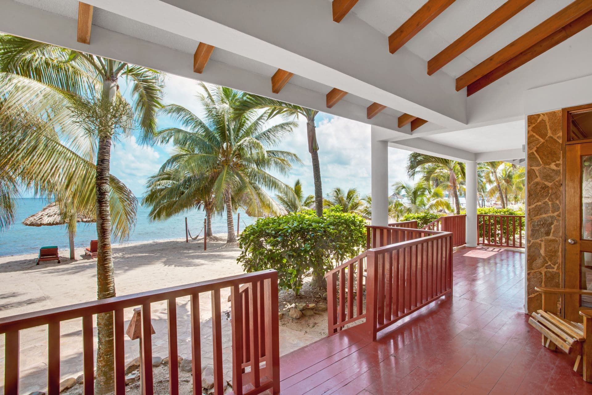 A spacious balcony with a wooden railing overlooking a sandy beach and the ocean. Palm trees line the beach, and there are umbrellas and lounge chairs visible. The sky is blue and sunny.