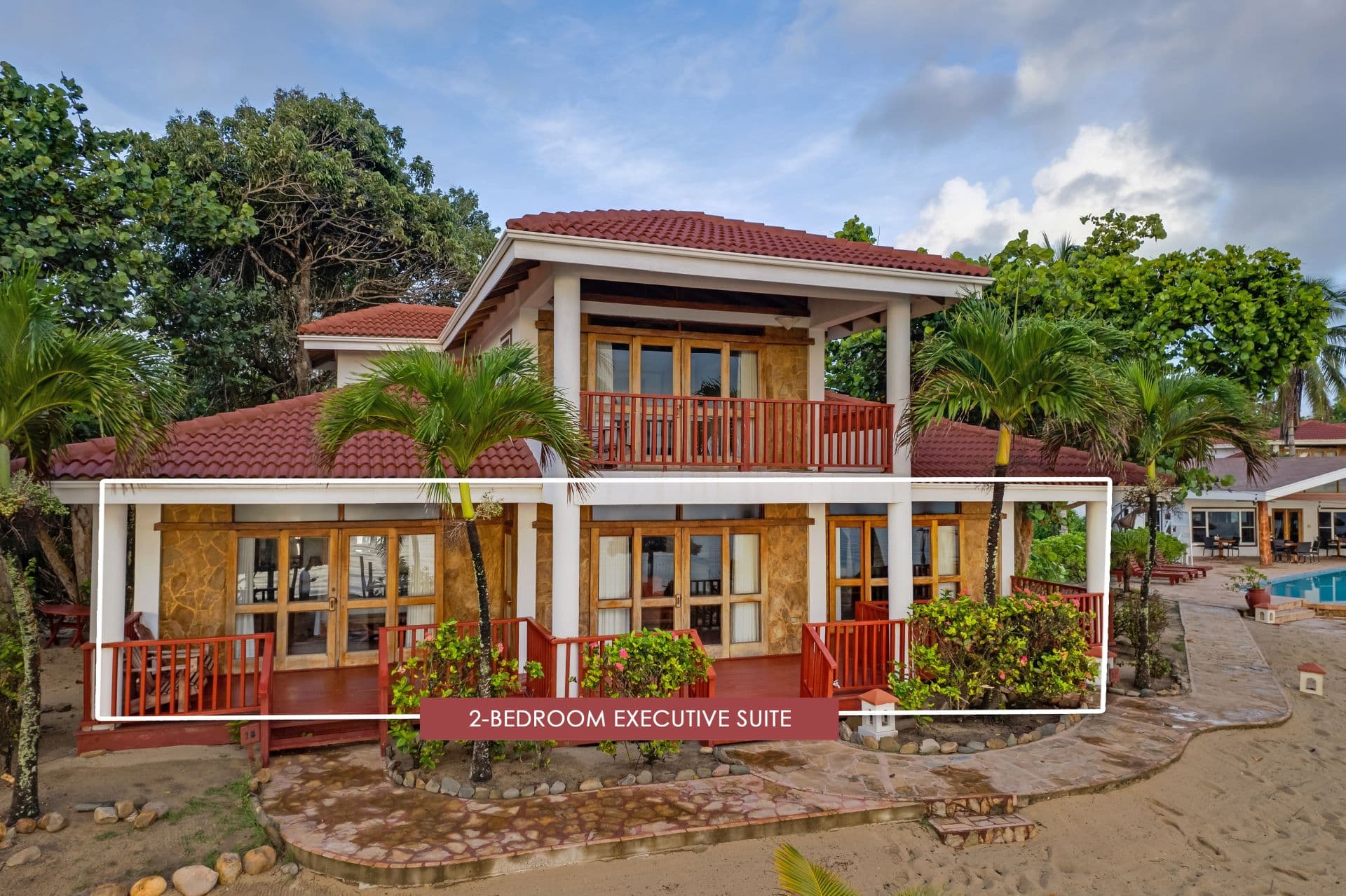 The image shows a 2-bedroom executive suite at Belizean Dreams Resort in Hopkins Bay, Belize. The suite is a two-story building with a red tile roof and a large balcony on the second floor. The suite is surrounded by palm trees and lush greenery.