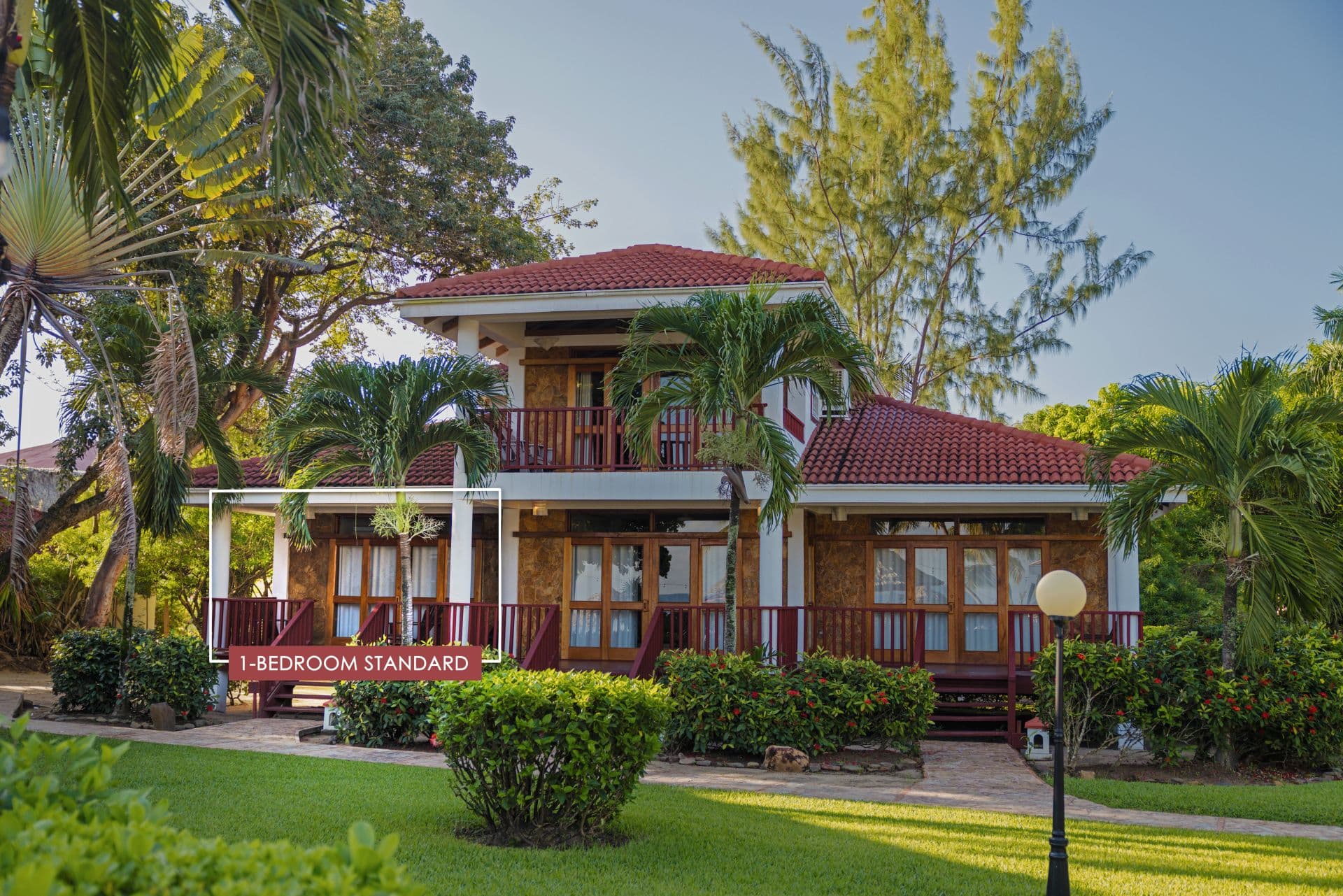 The image shows a 1-bedroom standard room at Belizean Dreams Resort in Hopkins Bay, Belize. The room is a two-story building with a red tile roof and a large balcony on the second floor. The room is surrounded by palm trees and lush greenery.