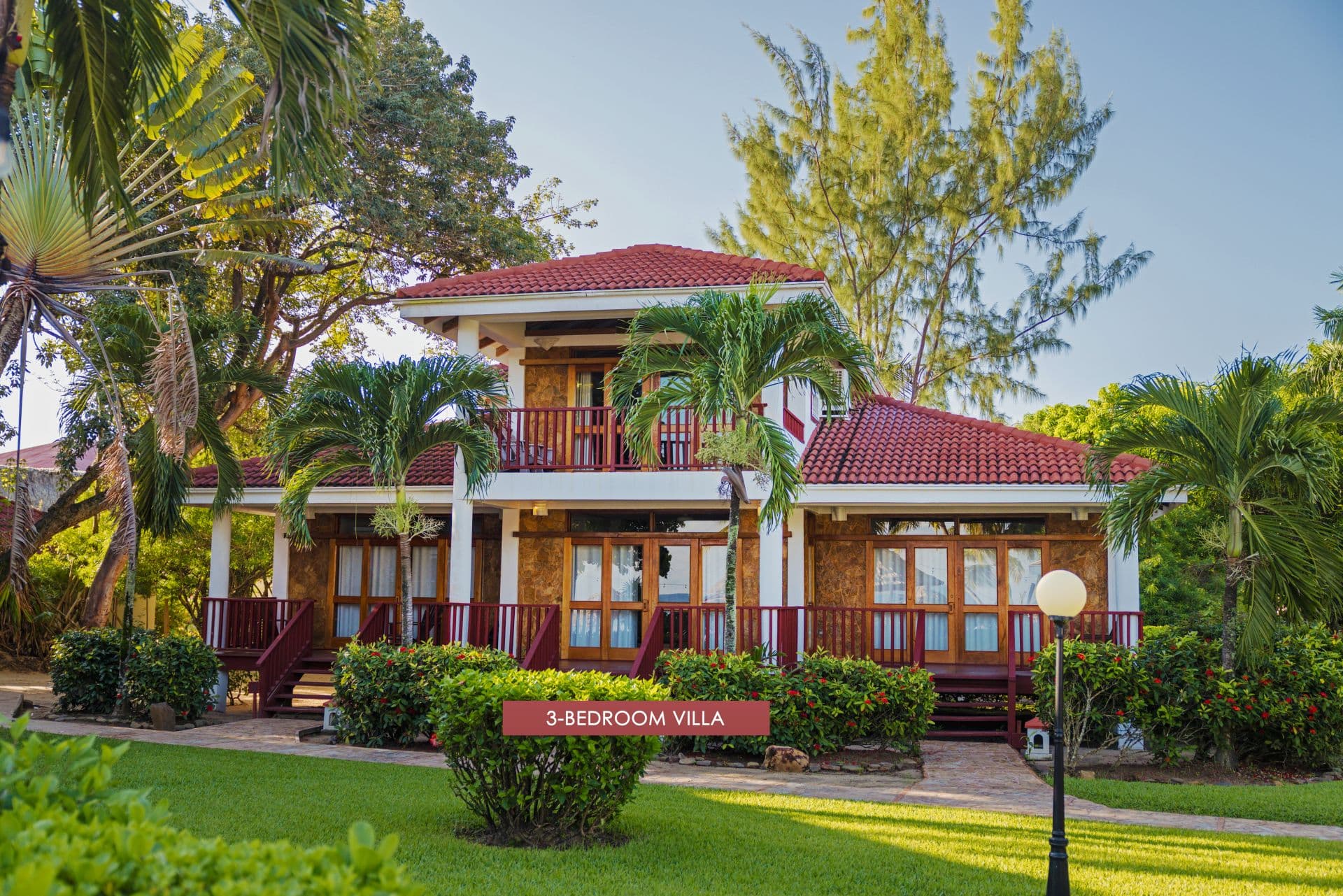 The image shows a 3-bedroom villa at Belizean Dreams Resort in Hopkins, Belize. The villa is a two-story building with a red tile roof and a large balcony on the second floor. The villa is surrounded by palm trees and lush greenery.