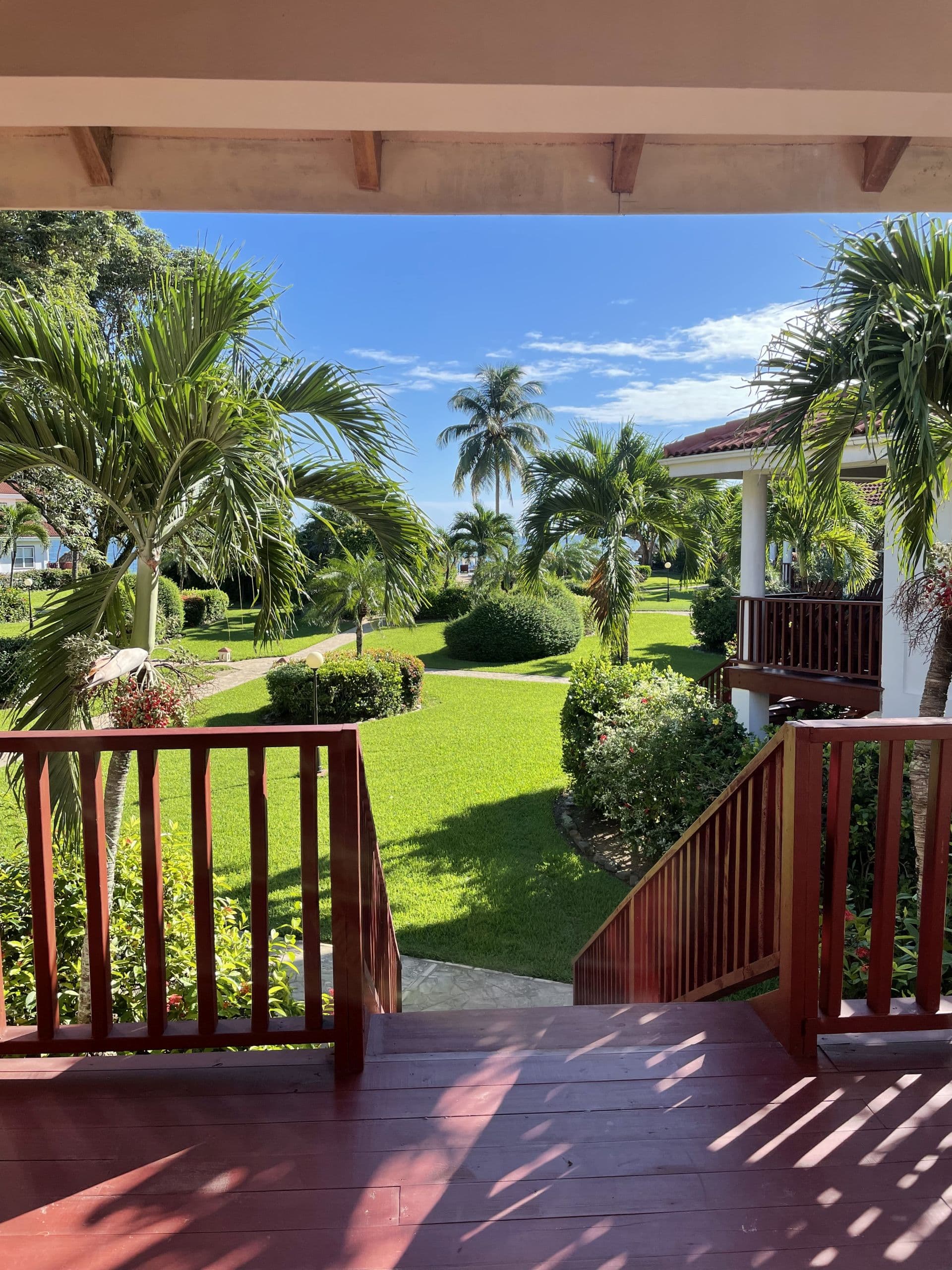 A wooden balcony overlooking a lush green lawn with palm trees. The ocean can be seen in the background, and the sky is blue and sunny.