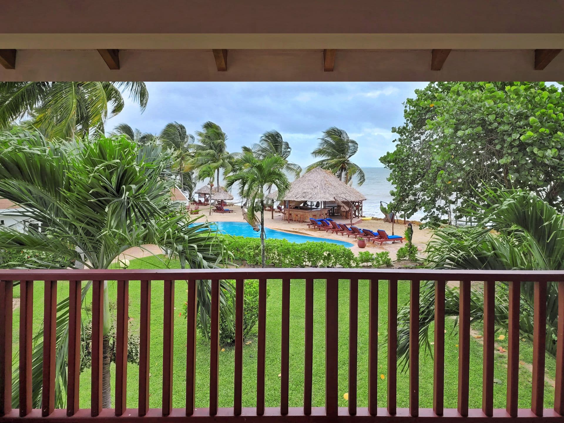 A balcony overlooking a tropical resort. There is a swimming pool, a bar, and palm trees in the foreground. The ocean is visible in the background, and the sky is cloudy.