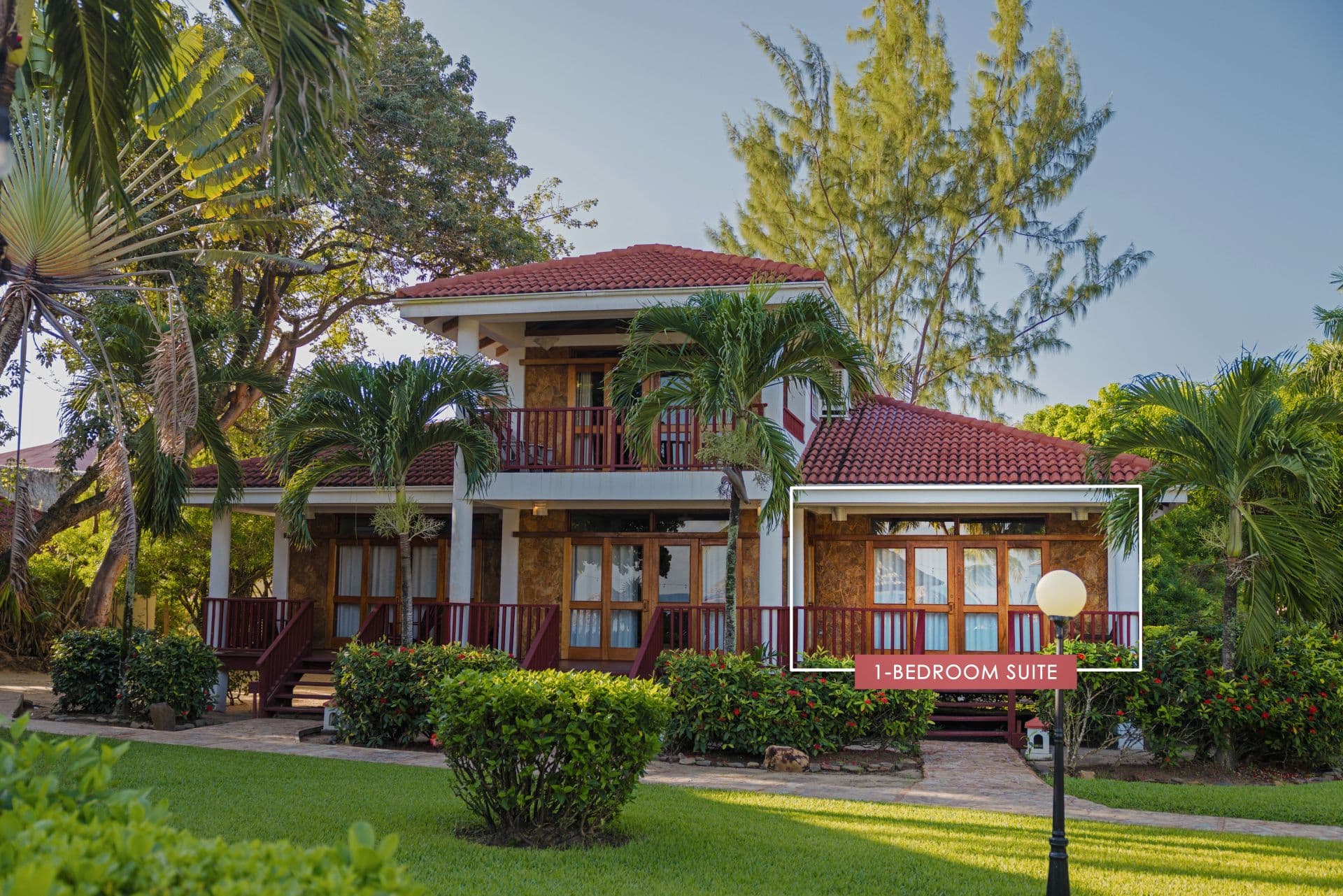 The image shows a 1-bedroom suite at Belizean Dreams Resort in Hopkins Bay, Belize. The suite is a two-story building with a red tile roof and a large balcony on the second floor. The suite is surrounded by palm trees and lush greenery.