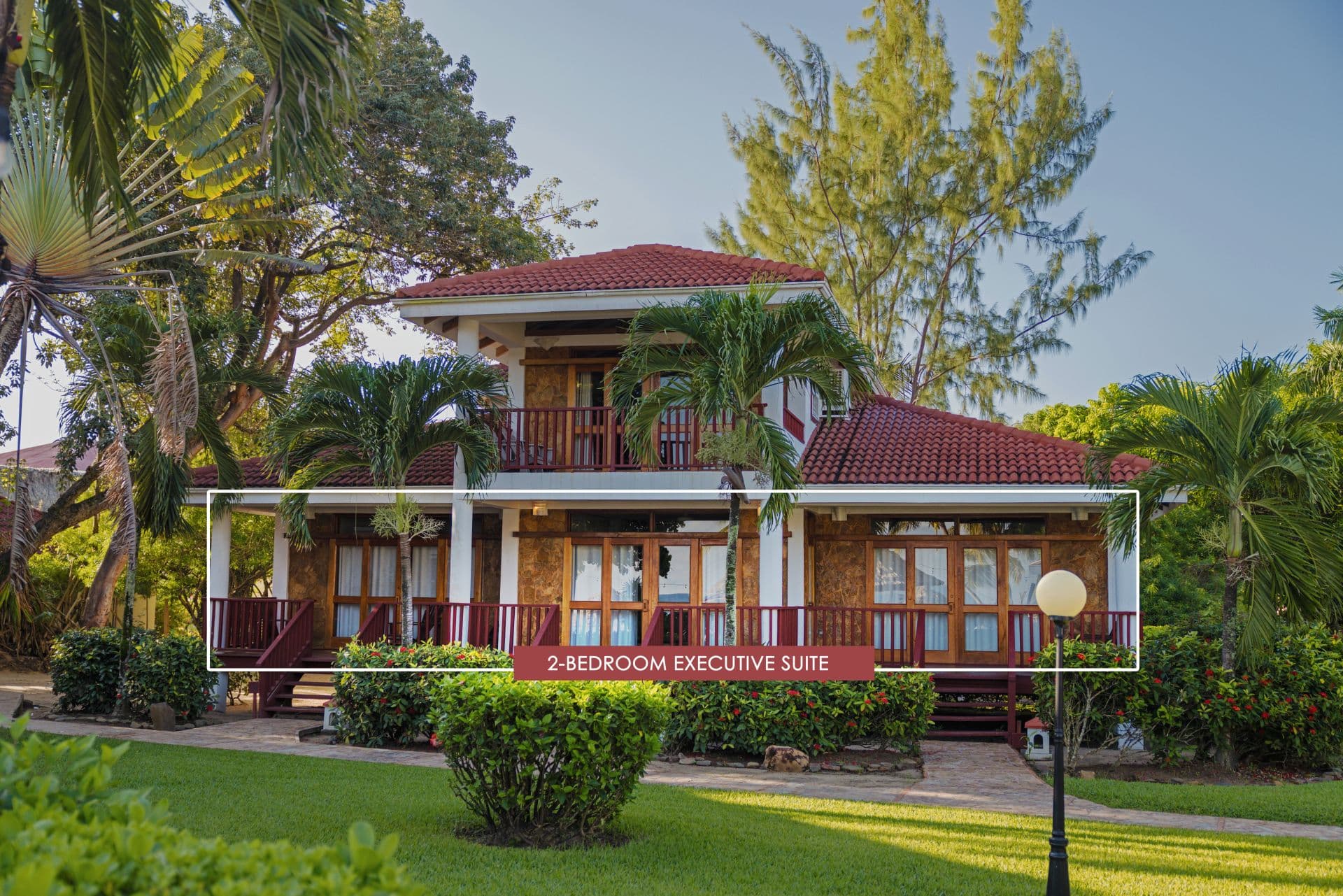 The image shows a 2-bedroom executive suite at Belizean Dreams Resort in Hopkins Bay, Belize. The suite is a two-story building with a red tile roof and a large balcony on the second floor. The suite is surrounded by palm trees and lush greenery.