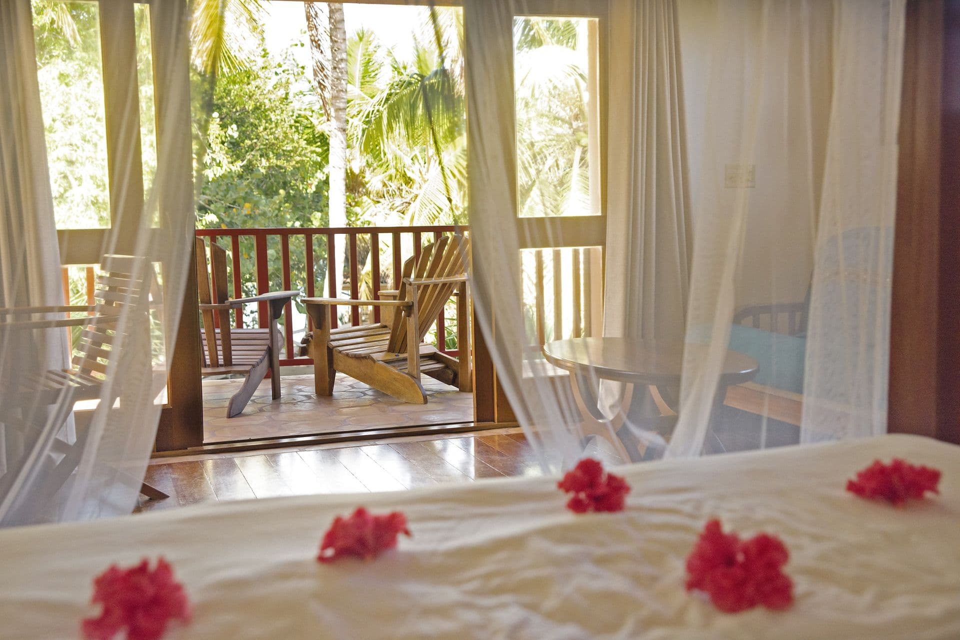 A bedroom with a large bed covered in white sheets and adorned with red flowers. Sheer white curtains frame a sliding glass door leading to a private balcony overlooking a lush tropical garden. Two wooden chairs and a small table are visible on the balcony. The room has a warm and inviting atmosphere with wooden floors and natural light.