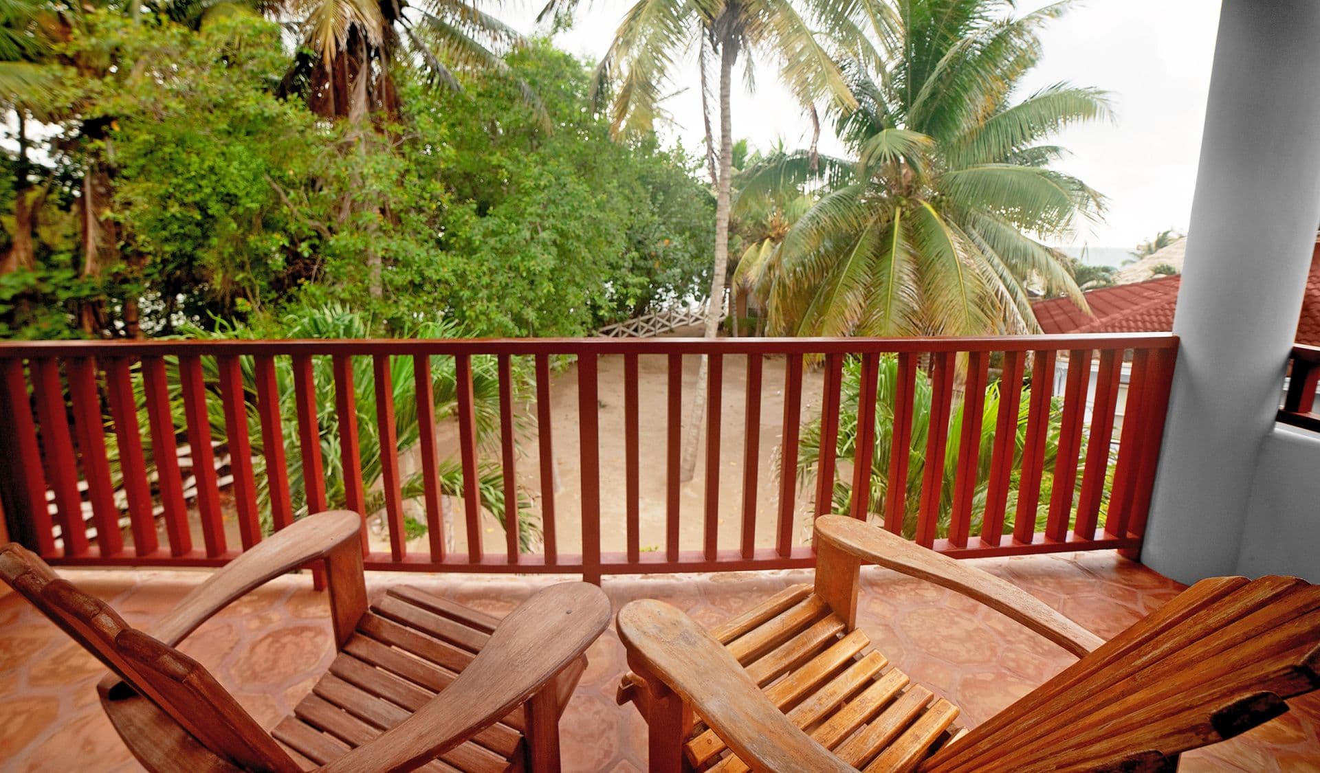 A balcony with two wooden chairs overlooking a tropical landscape. Tall palm trees dominate the scene, and lush greenery fills the background. The railing of the balcony is painted red, and there is a hint of a beach in the distance.