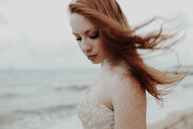 A red-haired woman in a beaded dress gazes thoughtfully at the ocean, her hair flowing in the breeze.