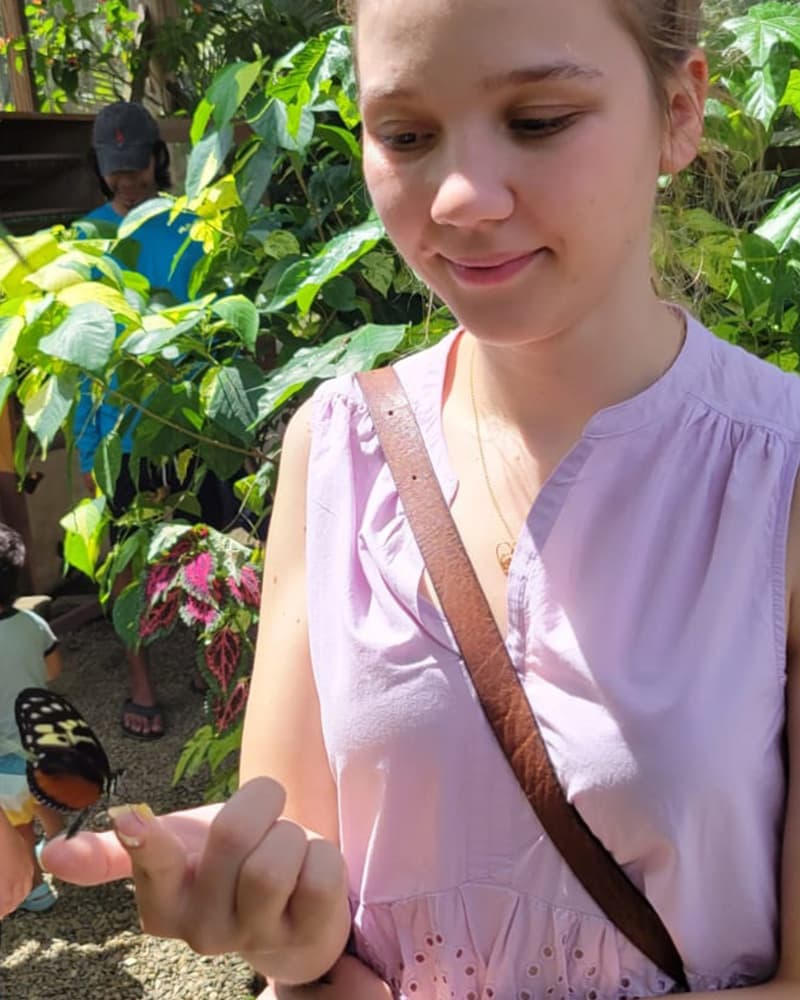 A young woman in a light purple top gently holds a butterfly on her hand amidst lush greenery.