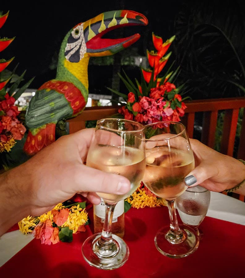 Two glasses of white wine are toasting in front of colorful tropical decorations and a carved parrot.
