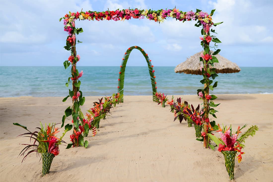 Tropical beach wedding ceremony setup with palm leaf arches, colorful flowers, and a walkway leading to the ocean