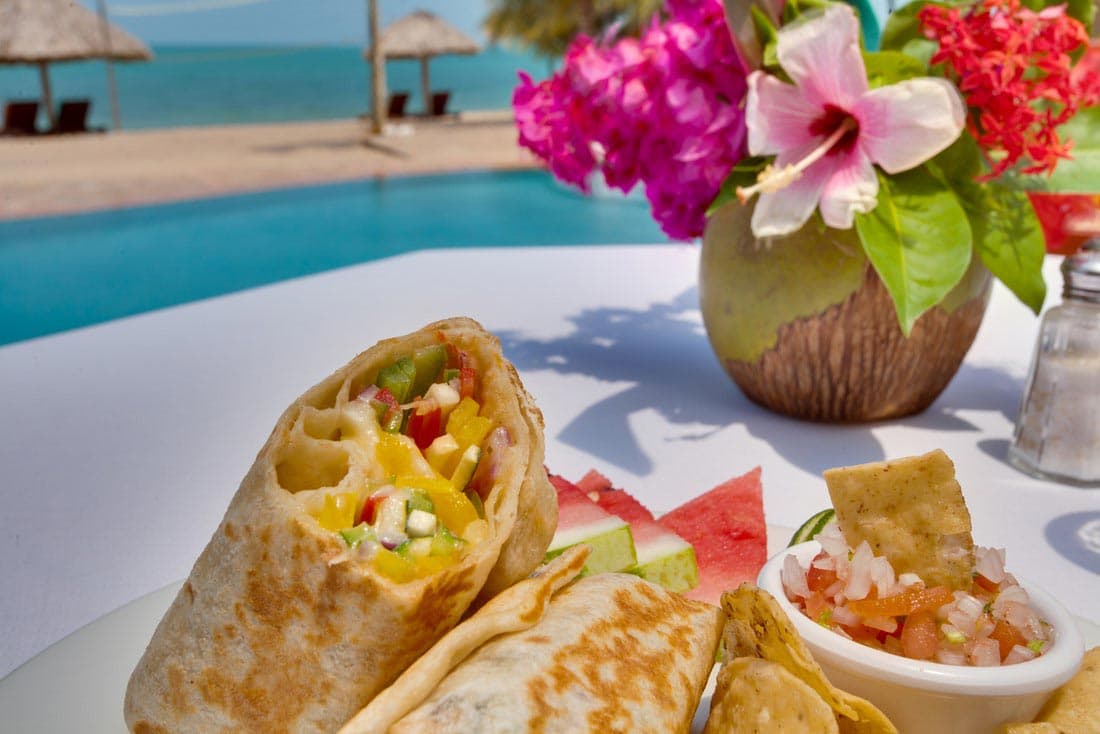 A delicious-looking seafood eggroll filled with grilled vegetables is served on a white plate alongside a bowl of salsa and a slice of watermelon. The background shows a pool and beach at a tropical resort.