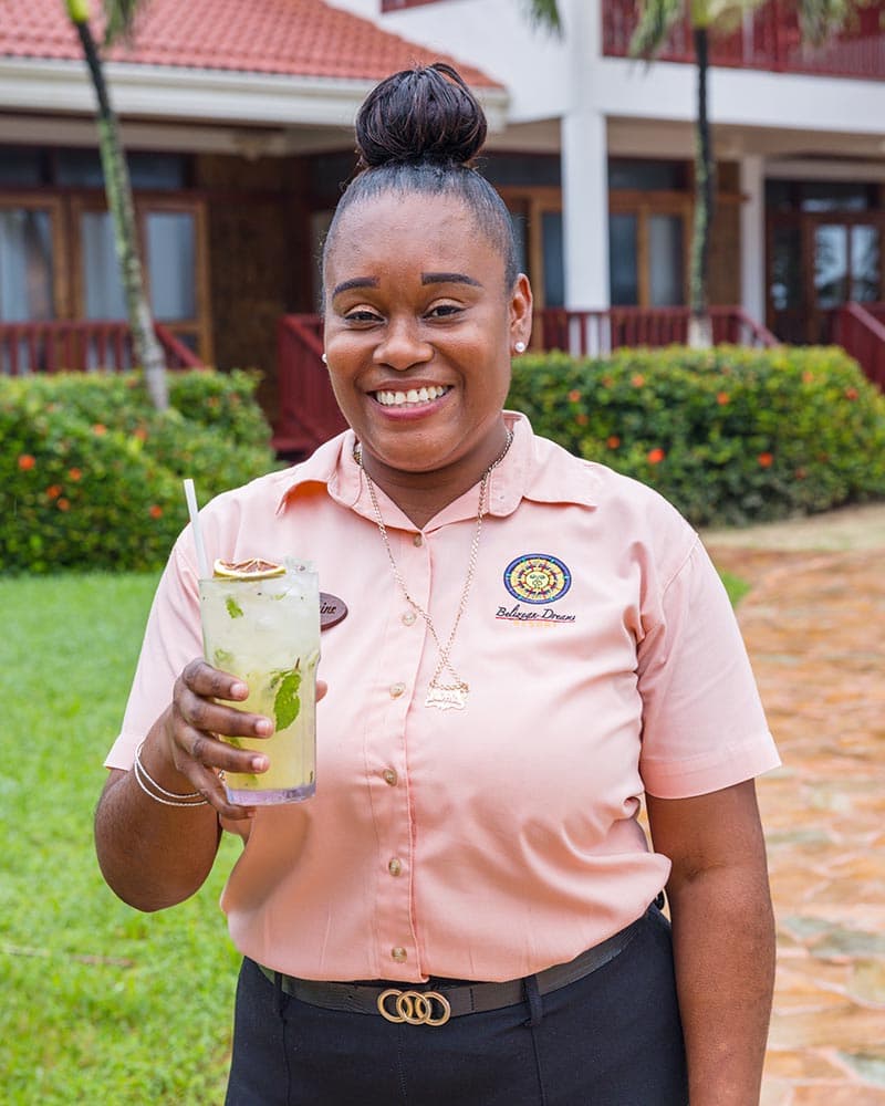 A smiling woman in a pink shirt holds a refreshing drink in a tropical setting.