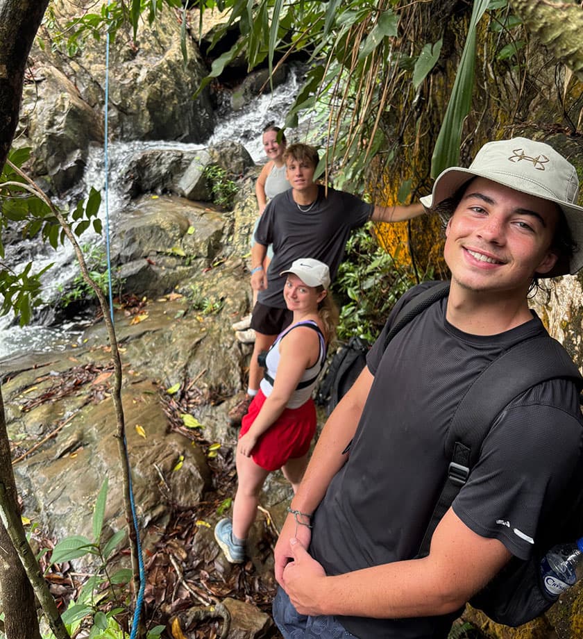 A group of four friends poses beside a small waterfall in a lush, green landscape.