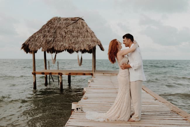 A couple embraces on a pier over the ocean, with a thatched-roof gazebo in the background.