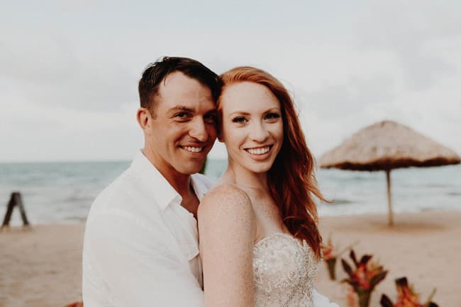 A smiling couple embraces on a beach, with a thatched umbrella in the background.