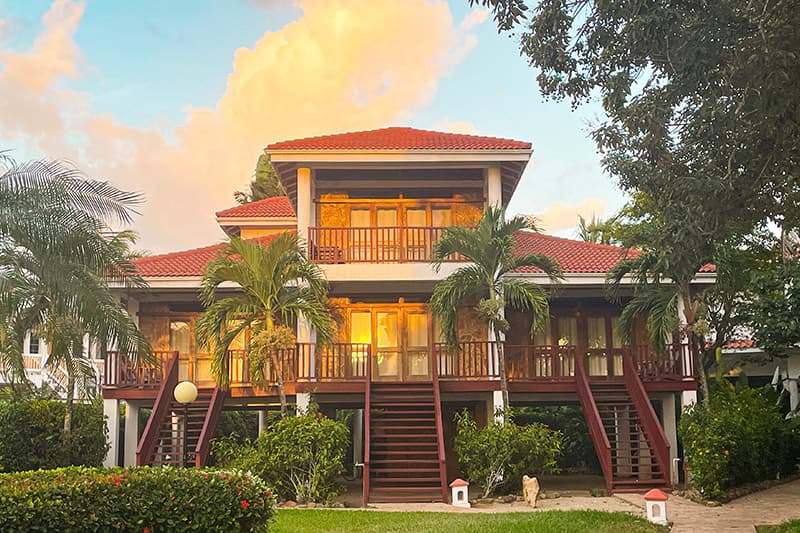 A large two-story house with a red roof surrounded by greenery and palm trees at sunset. A large two-story house with a red roof surrounded by greenery and palm trees at sunset.
