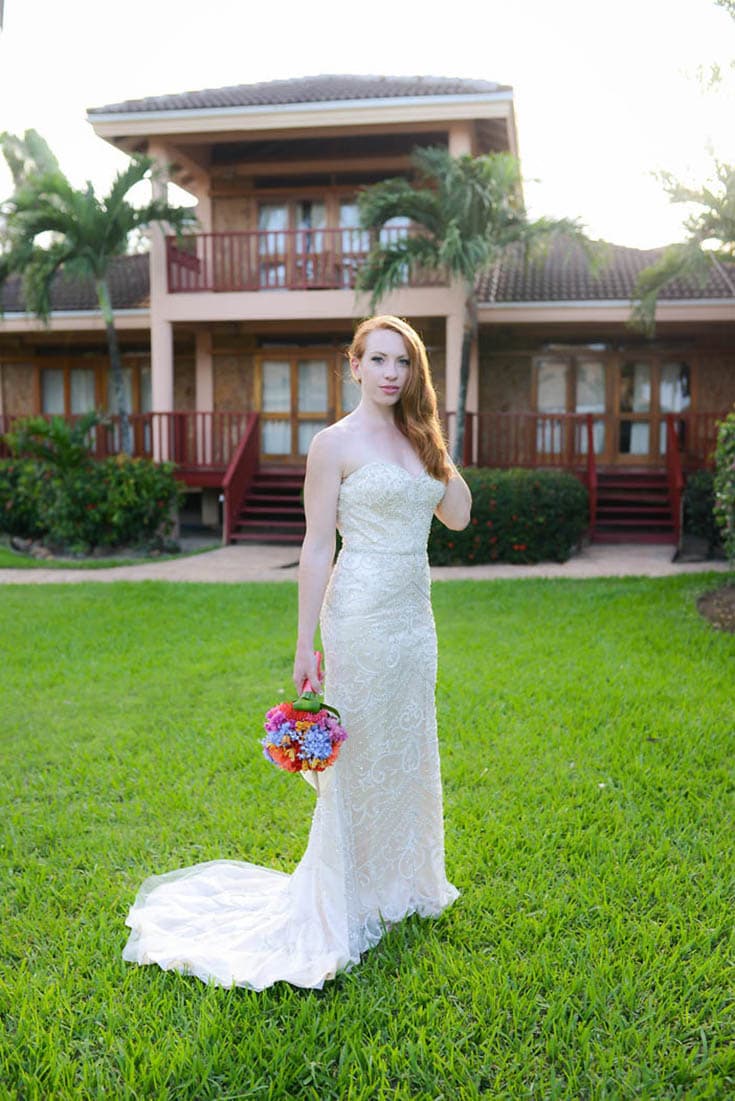 A beautiful auburn woman in a wedding gown poses on the lawn in front of a villa with a 2-story balcony.
