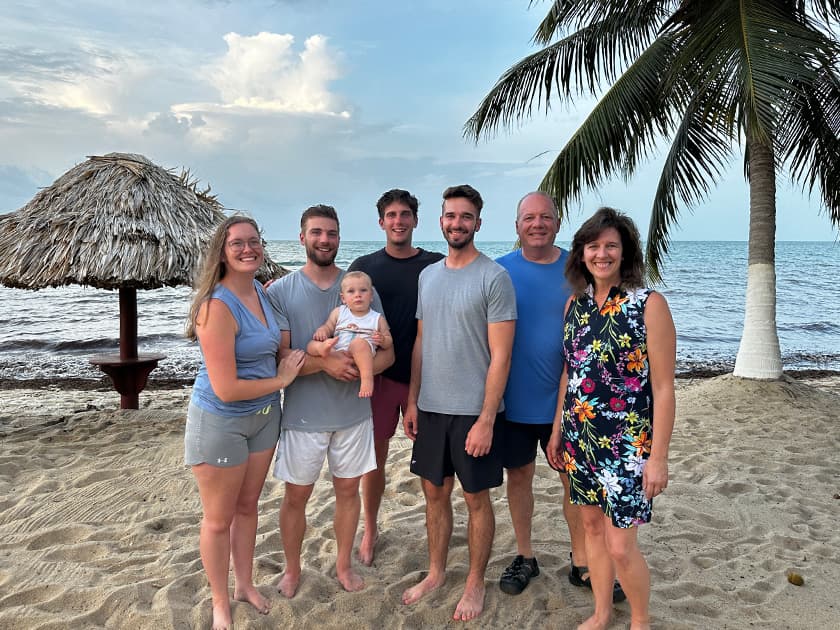 A group of eight people, including a baby, pose together on a sandy beach near the ocean. A group of eight people, including a baby, pose together on a sandy beach near the ocean.