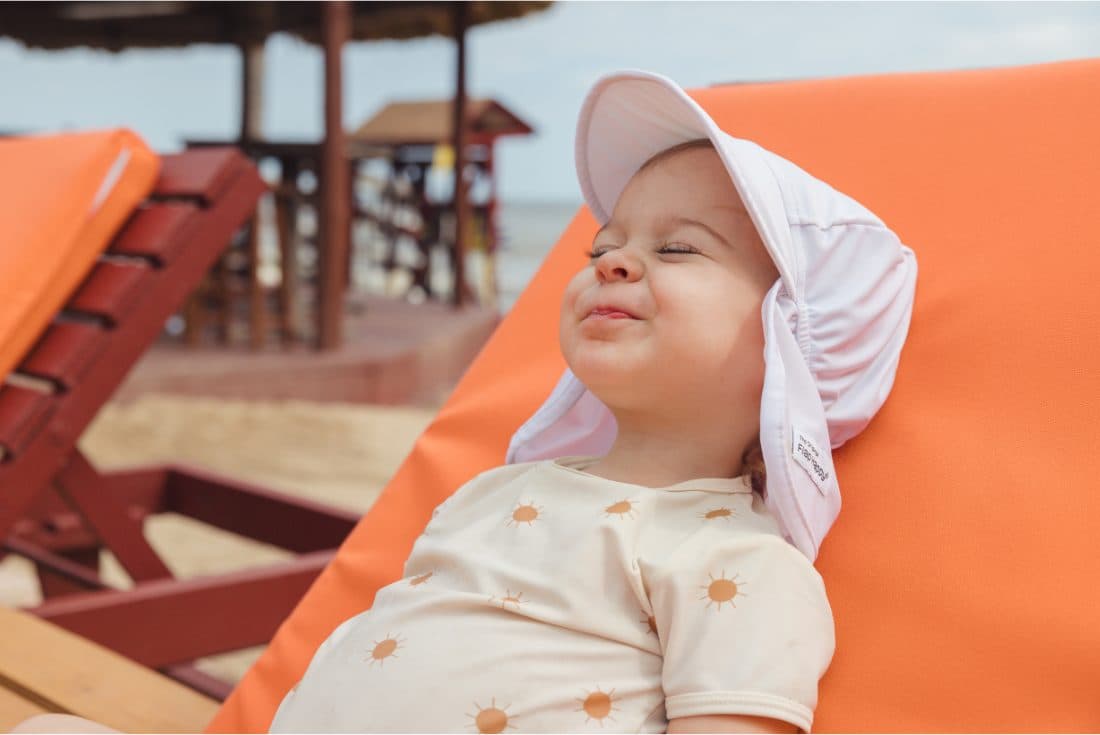Cute toddler in a white sun hat rests smiling on a cabana chair on the beach.