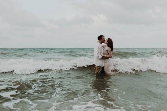 A couple embraces in the ocean, standing in the water as waves crash around them.