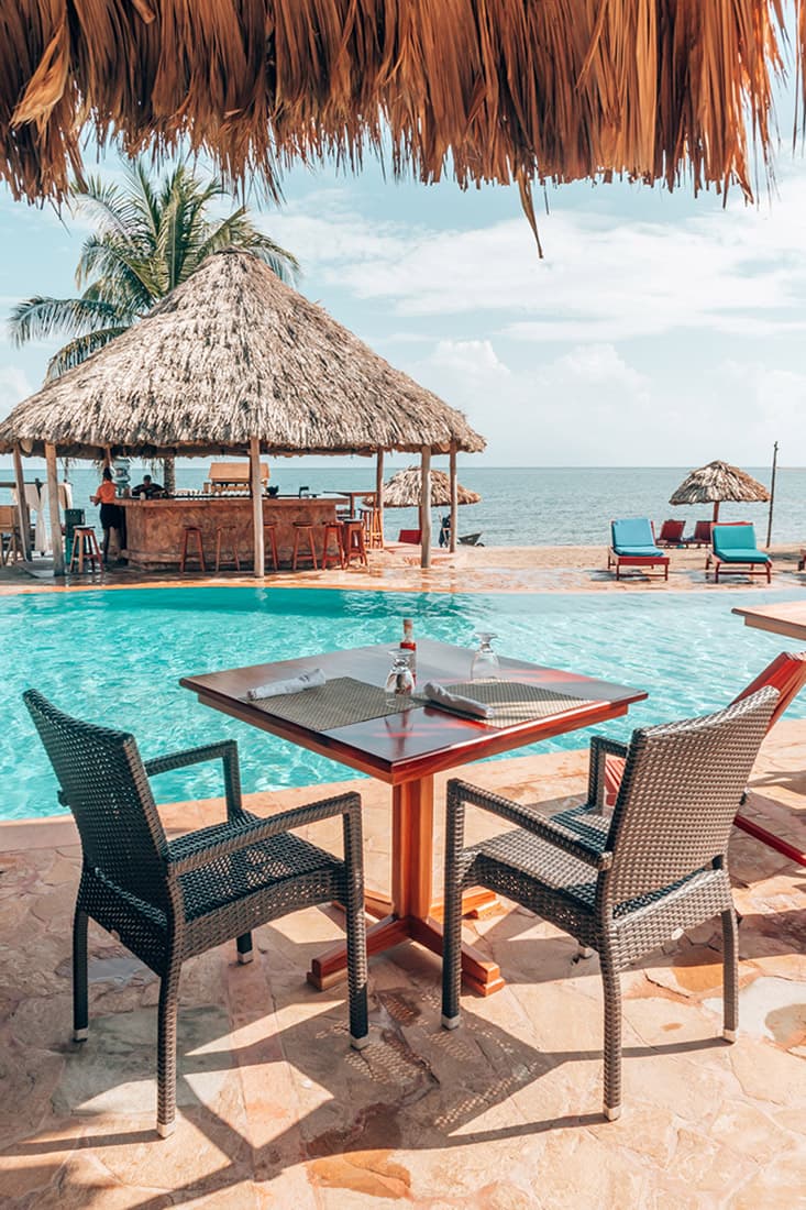 A picturesque beachfront scene at Belizean Dreams Resort. A thatched-roof bar overlooks a swimming pool and the ocean. Lounge chairs are arranged around the pool, and a wooden table with chairs is set up for outdoor dining. The sky is clear and blue.