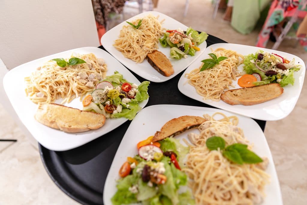 Four plates of food on a black tray. Each plate contains a portion of spaghetti with a creamy sauce, a side salad with mixed greens and vegetables, and a piece of toasted bread. The plates are arranged in a square formation.