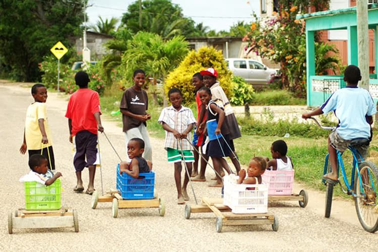 A group of children pull toy carts with toddlers along a rural road.