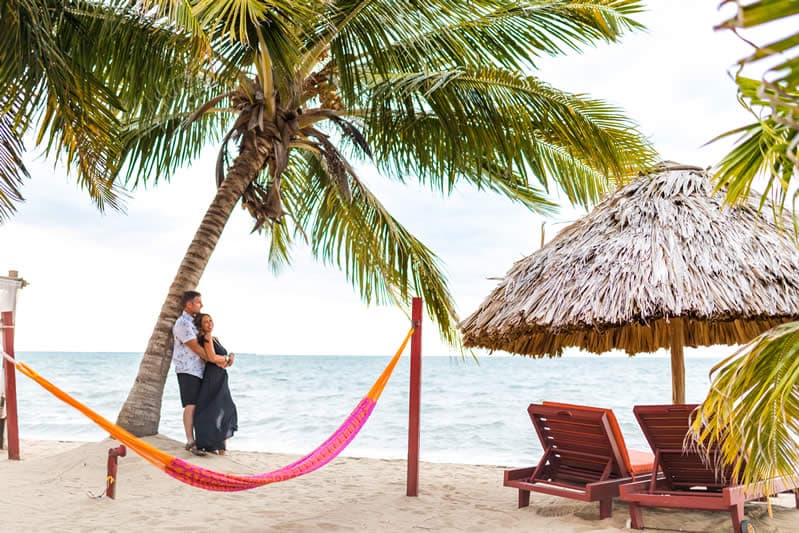 A couple embraces on a beach under palm trees, with a hammock and thatched-roof hut in the background.
