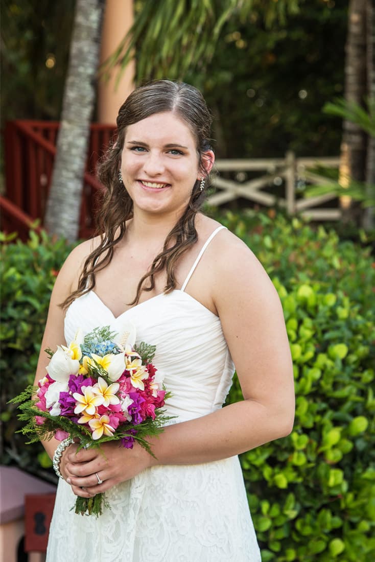 Pretty young bride in a white dress poses in front of hedges, holding a colorful wedding bouquet.