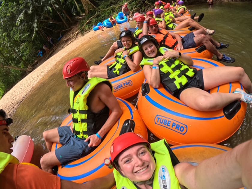 A group of people in orange inflatable tubes wearing life jackets smiles for a selfie while floating on a river.