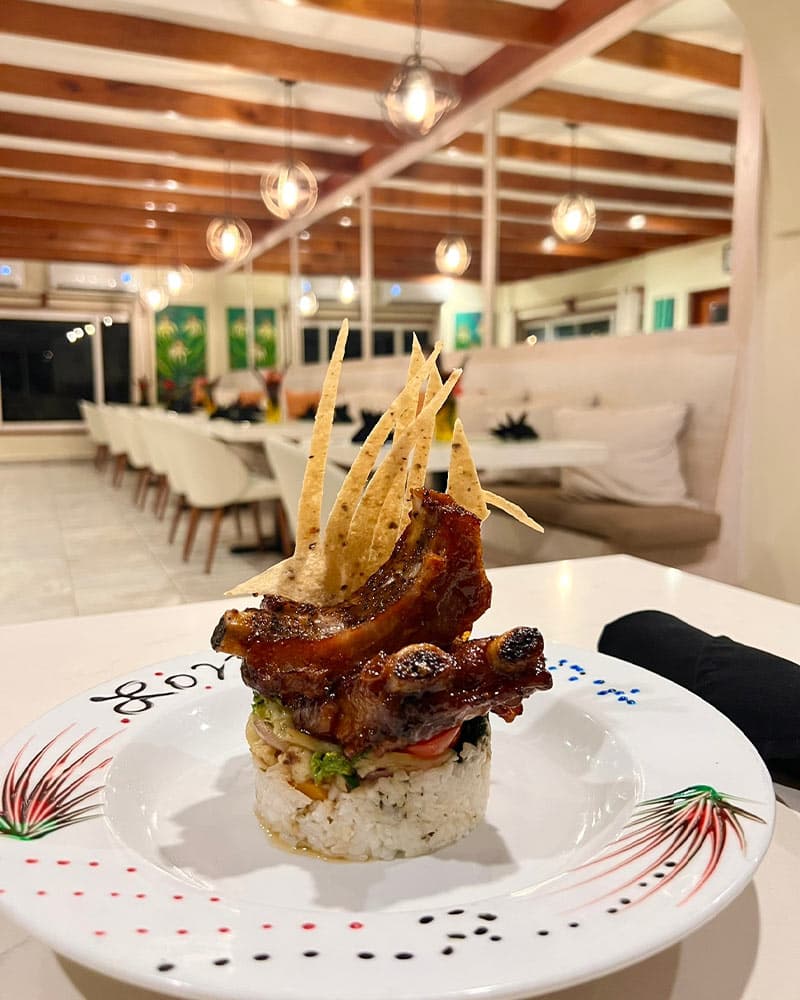 A gourmet dish featuring pork ribs served on a bed of rice. The ribs are topped with a colorful array of vegetables and crispy tortilla chips. The dish is presented on a white plate with a decorative design. The restaurant's interior is visible in the background, with white tablecloths and hanging lights.