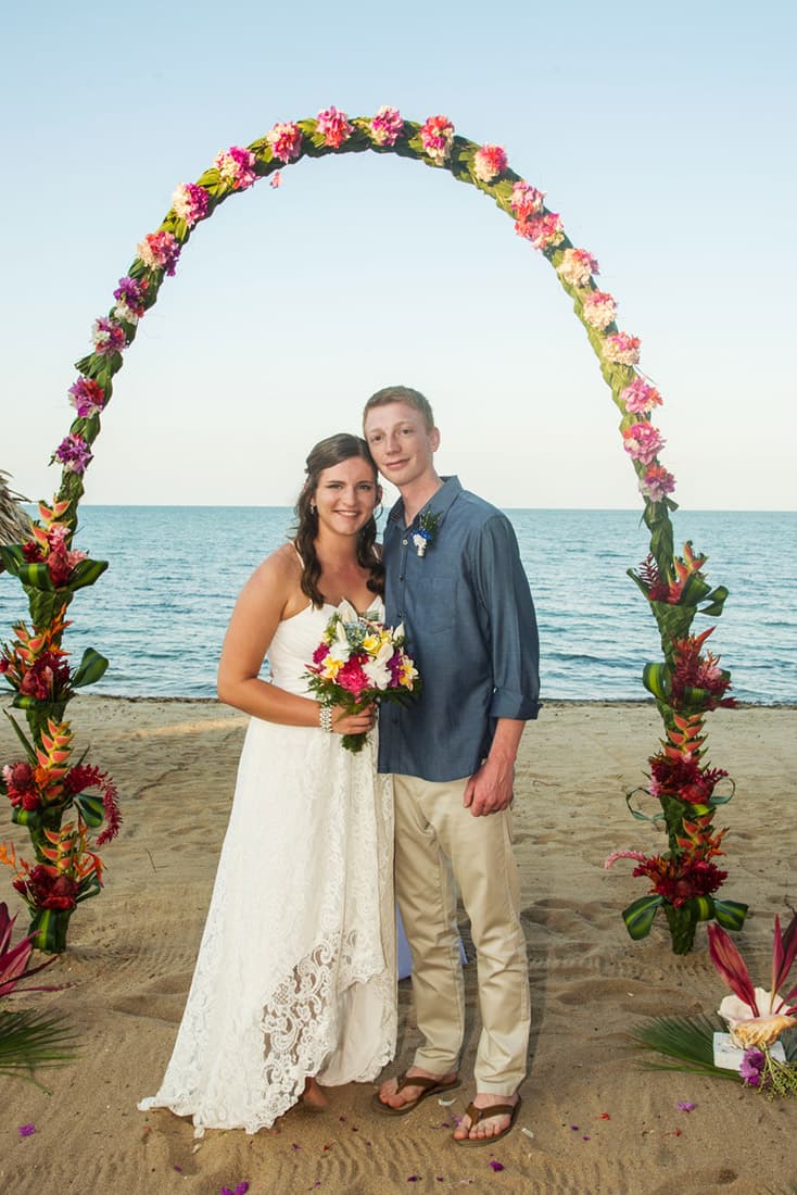 Young bride and groom pose on the beach under a wedding arch decorated with pink, green and orange tropical flowers.
