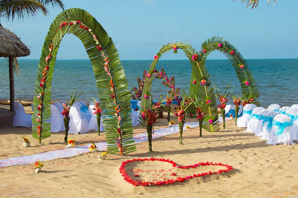 A beach wedding ceremony setup with palm leaf arches decorated with flowers. White chairs are arranged in rows facing the ocean. A heart shape is drawn in the sand with red petals.